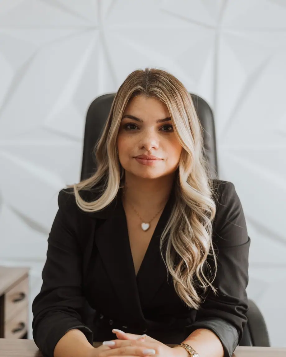 Dra. Camila Chagas sentada à mesa do escritório, de blazer preto, mãos entrelaçadas
