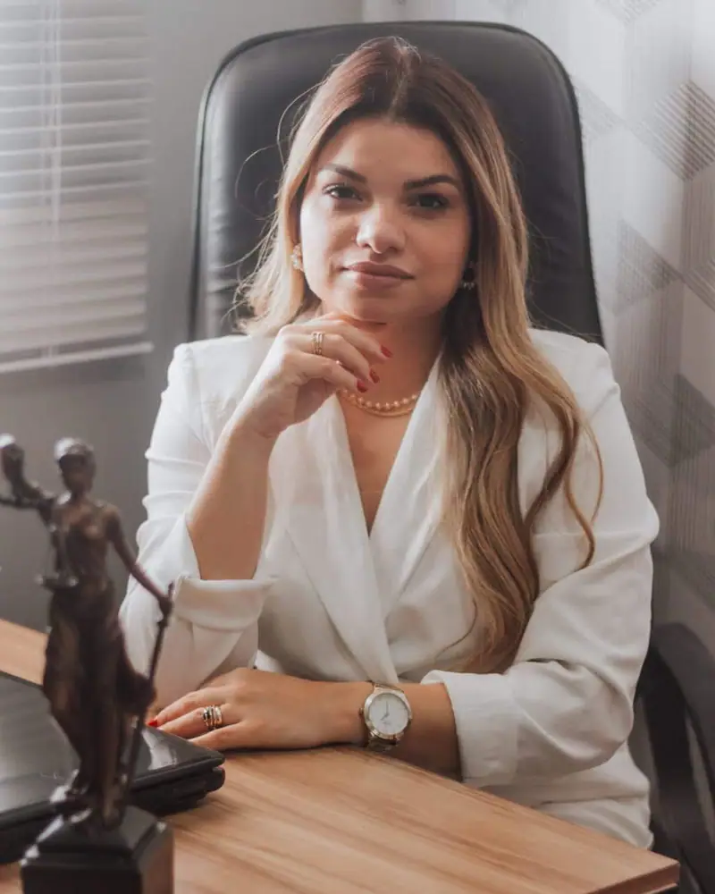 Dra. Camila Chagas à mesa do escritório, de blazer branco, com a estatueta da Justiça
