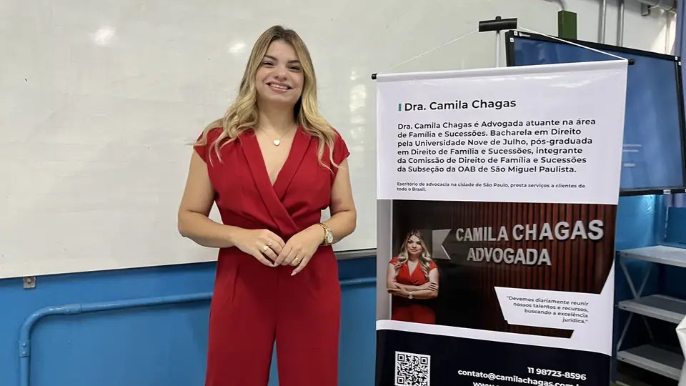 Dra. Camila Chagas em pé numa sala de aula, ao lado de um banner com o nome e a apresentação dela